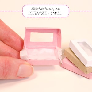 May include: A miniature pink bakery box with a clear window, held by a hand. The box is open, revealing white tissue paper inside. The text "Miniature Bakery Box RECTANGLE - SMALL" is above the box. Other boxes are in the background.