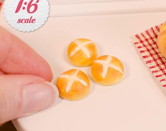 May include: Three miniature golden-brown round buns, each featuring a white cross pattern on top. The items are labeled as 1:6 scale. These small, baked-style goods are presented on a smooth, light-colored surface next to a red and white checkered cloth.