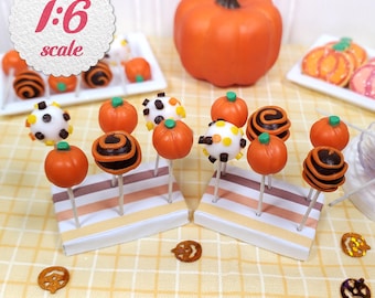 May include: Miniature Halloween-themed cake pops and a pumpkin. The cake pops are orange, white, and brown, with various designs. The image includes the text "1:6 scale" in a circular design. The background is a yellow checkered pattern.