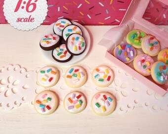 May include: A collection of miniature 1:6 scale cookies with white icing and colorful sprinkles. The set includes round cookies on a small white plate, loose cookies arranged on a white lace mat, and cookies inside a pink cardboard box.
