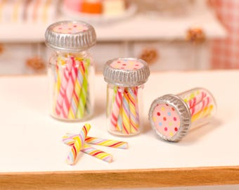 May include: Miniature glass jars with silver lids, filled with colorful candy canes. The jars are clear, showcasing the striped candies inside. Some candy canes are scattered nearby, creating a whimsical scene. The lids are decorated with pink and colorful polka dots.