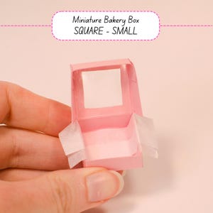 May include: A small, pink, square miniature bakery box with a clear window on the top. The box is open, revealing white tissue paper inside. The text "Miniature Bakery Box SQUARE - SMALL" is above the box.