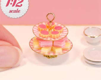 May include: A miniature two-tiered dessert stand featuring pink and white striped plates, holding small pink and yellow cakes. The stand has a gold-colored metal handle and base. A white circular label in the corner displays the text 1:12 scale.