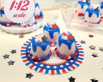 May include: Miniature desserts with white frosting, blue icing drips, and red, white, and blue accents. Each cake is topped with a star. The image includes a decorative plate and star-shaped confetti. The text "1:12 scale" is visible.