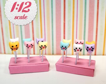 May include: A collection of miniature 1:12 scale cat-shaped lollipops in various pastel colors, each featuring a painted face with whiskers and a small heart nose. The lollipops are displayed upright in two rectangular pink foam blocks.