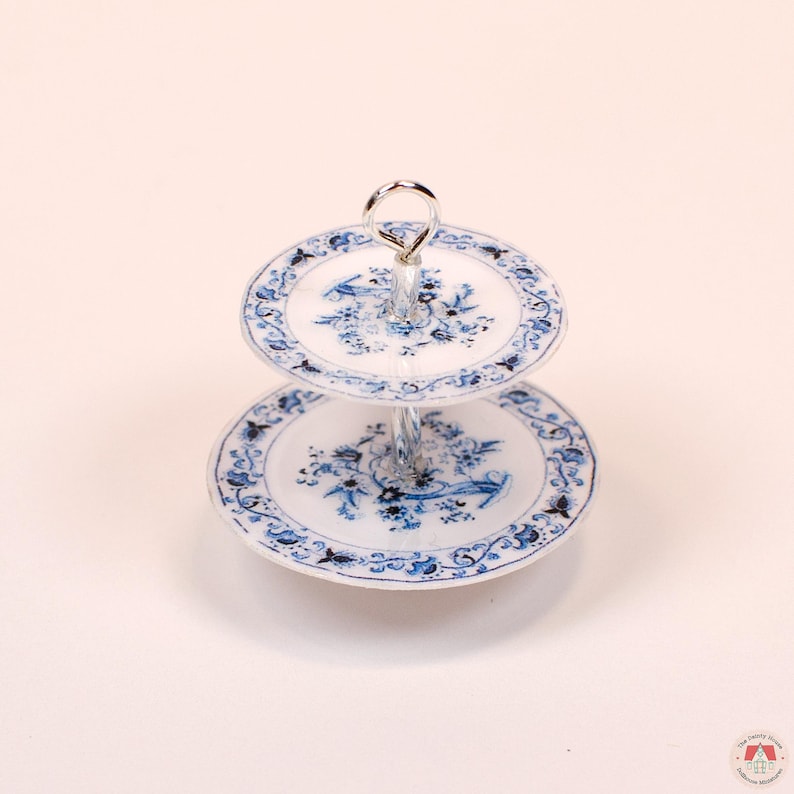 Puede incluir: Un soporte de servicio de dos niveles con platos de cer&aacute;mica blanca decorados con intrincados patrones florales azules. Los niveles est&aacute;n conectados por un poste central de metal plateado que termina en un asa circular en la parte superior.