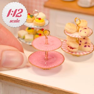 Puede incluir: Un soporte de servicio en miniatura de dos niveles con acabado rosa brillante, bordes dorados y un asa central en forma de anillo. El soporte cuenta con dos platos redondos con una textura estriada sutil, dise&ntilde;ado a escala 1:12 para casas de mu&ntilde;ecas.