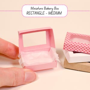 May include: A miniature pink bakery box with a clear window, held by a hand. The box is open, revealing white tissue paper inside. The text "Miniature Bakery Box RECTANGLE - MEDIUM" is above the box.