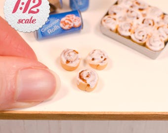 May include: A 1:12 scale miniature set featuring three small cinnamon rolls with white icing, a rectangular metal tray filled with twelve baked cinnamon rolls, and two blue cylindrical containers labeled Cinnamon Rolls.