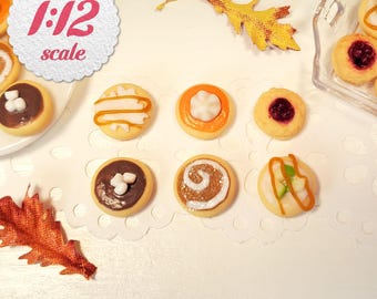 May include: A collection of miniature 1:12 scale cookies with various toppings including chocolate, marshmallow, fruit jam, and icing swirls. The set includes round cookies with orange, brown, and white decorations, arranged with two decorative autumn leaves.