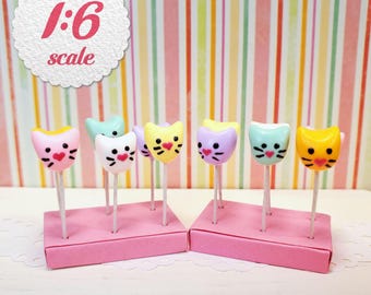 May include: A collection of miniature cat-shaped lollipops in various pastel colors, each featuring a heart-shaped nose and whiskers, displayed on thin white sticks in two pink rectangular stands, with a 1:6 scale label visible.