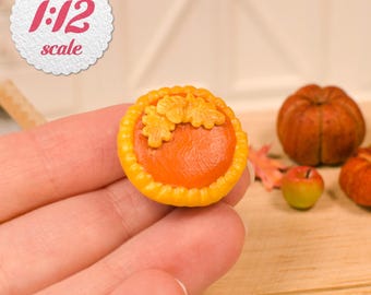 May include: Miniature pumpkin pie with a bright orange filling and a yellow crust, topped with two leaf-shaped decorations. The pie is held in a hand, with a pumpkin and apple in the background. The image includes a label that says "1:12 scale".