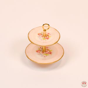 Puede incluir: Un soporte de dos niveles con dos platos redondos de color rosa p&aacute;lido con un estampado floral de rosas rosas y hojas verdes. Los platos tienen bordes pintados en oro y est&aacute;n unidos por un poste central de metal dorado con un asa de anillo.