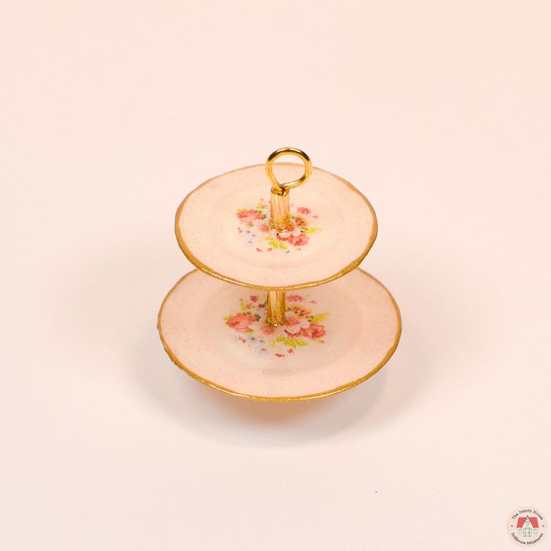 Puede incluir: Un soporte de dos niveles con dos platos redondos de color rosa p&aacute;lido con un estampado floral de rosas rosas y hojas verdes. Los platos tienen bordes pintados en oro y est&aacute;n unidos por un poste central de metal dorado con un asa de anillo.