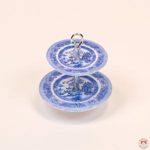 Puede incluir: Un soporte de dos niveles con platos de cer&aacute;mica azul y blanca con un patr&oacute;n tradicional de sauce. Los platos est&aacute;n conectados por una varilla central de metal plateado que se extiende hacia arriba formando una peque&ntilde;a asa circular.