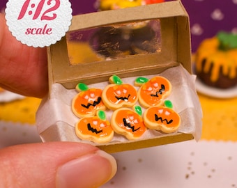 May include: A miniature 1:12 scale cardboard box containing six small orange pumpkin-shaped cookies with green stems and black hand-painted zigzag facial expressions. The box features a clear rectangular window and white parchment paper lining.