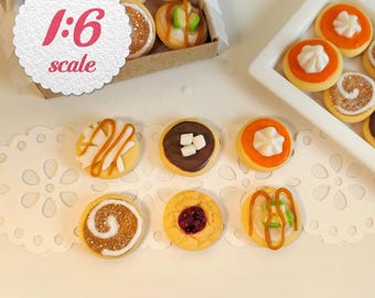 May include: A collection of miniature 1:6 scale cookies in a cardboard box and on a white tray. The cookies feature various toppings including white icing, chocolate, fruit jam, marshmallows, and caramel drizzles. The set includes round biscuits with diverse textures and colors, presented on a white lace-patterned paper doily.