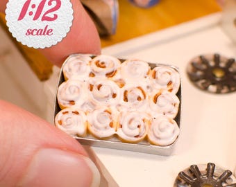 May include: A miniature rectangular metal baking pan containing twelve individual cinnamon rolls topped with white icing, presented in 1:12 scale. The rolls are arranged in a grid pattern, showing a swirled brown and white texture.