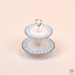 Puede incluir: Un soporte de dos niveles con platos circulares blancos y bordes de rayas azul claro. Los niveles est&aacute;n conectados por un poste central de metal plateado que se extiende hacia arriba formando un peque&ntilde;o bucle circular en la parte superior.