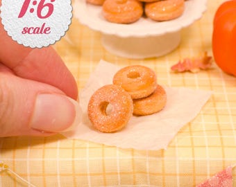 May include: A set of miniature 1:6 scale glazed donuts with a light brown color and a sugary texture. The donuts are arranged in a small stack on a piece of parchment paper, with additional donuts displayed on a white pedestal stand in the background.