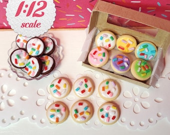 May include: A collection of miniature 1:12 scale cookies with white, pink, yellow, blue, purple, and green icing topped with colorful sprinkles. The cookies are arranged on a small clear plate, in a cardboard box, and on a white surface.