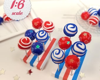 May include: Miniature red, white, and blue cake pops on sticks, arranged on a striped stand. Some pops have star-shaped sprinkles. The image includes the text "1:6 scale" in a circular frame.