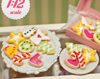 May include: Miniature Christmas cookies in various shapes, including stars, hearts, and trees, are arranged on a white plate and in a pink box. The cookies are decorated with colorful icing and are labeled as 1:12 scale.