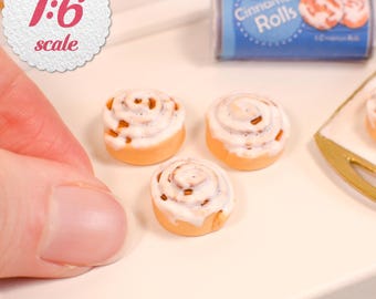 May include: Three miniature cinnamon rolls with white icing, presented in a 1:6 scale. A blue cylindrical package labeled Cinnamon Rolls is visible in the background, alongside a gold-toned tray edge, all on a smooth white surface.