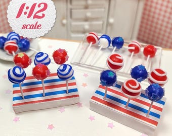 May include: Miniature cake pops in red, white, and blue, displayed on striped stands. The image includes a 1:12 scale label, suggesting a dollhouse or miniature food theme. The pops have various designs, including swirls and star sprinkles.