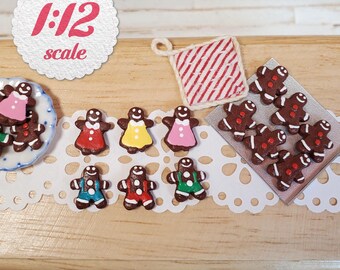 May include: A collection of miniature 1:12 scale gingerbread cookies, including figures wearing colorful icing outfits, a square red and white striped potholder, and a silver baking sheet, all arranged on a white decorative paper doily.