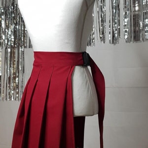 May include: A red pleated skirt with a black adjustable buckle belt. The skirt has a side slit and a long tie that can be worn as a belt.