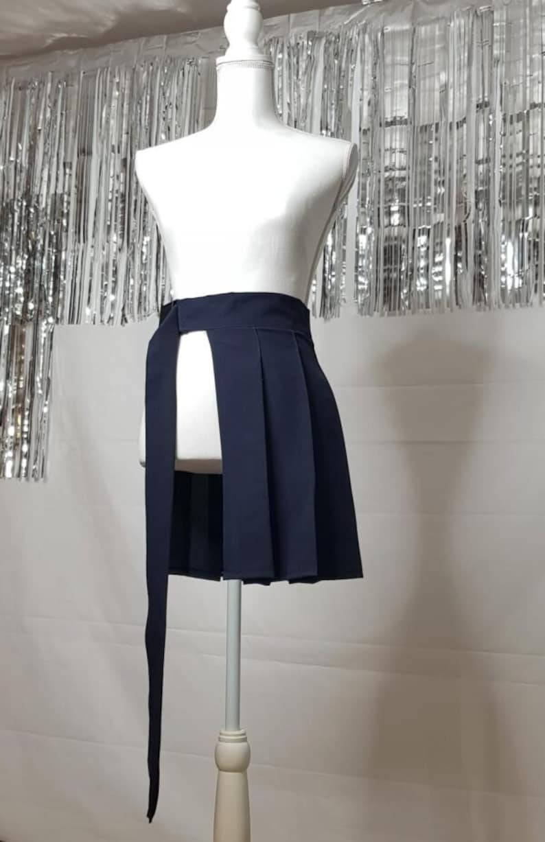 May include: A navy blue pleated skirt with a long tie that wraps around the waist. The skirt has a slit in the front and is displayed on a white mannequin.