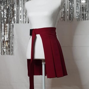 May include: A red pleated skirt with a black belt and a long tie. The skirt is on a white mannequin.