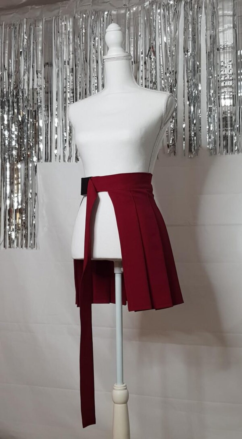 May include: A red pleated skirt with a black belt and a long tie. The skirt is on a white mannequin.