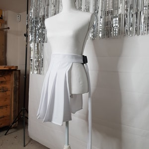 May include: White pleated skirt with a black belt and a long white tie. The skirt is on a white mannequin against a silver background.