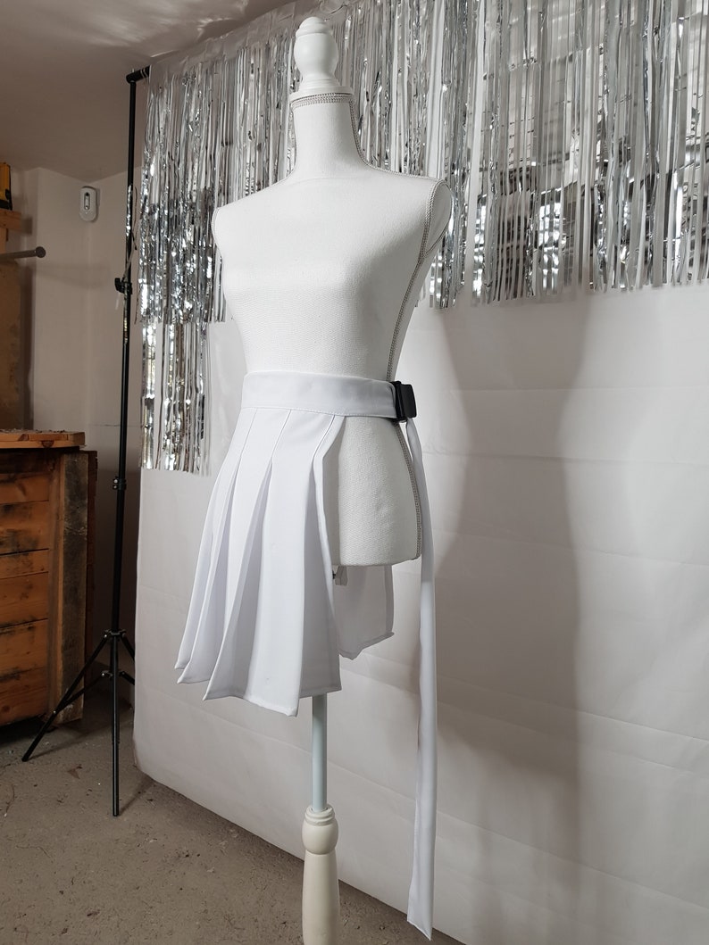 May include: White pleated skirt with a black belt and a long white tie. The skirt is on a white mannequin against a silver background.