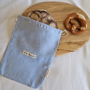 Könnte beinhalten: Hellblaue Brotbeutel-Tasche aus Leinen mit dem Text "Brotbeutel" auf einem Holzbrett. Ein Brotlaib und eine Brezel befinden sich ebenfalls auf dem Brett. Die Tasche hat einen Kordelzug.