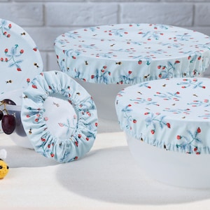 Può includere: Quattro coperchi per ciotole riutilizzabili con un motivo floreale blu e bianco con bacche rosse e api. I coperchi sono in tessuto e hanno un bordo arricciato. Una piccola ape all'uncinetto è in primo piano.