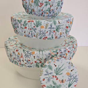 May include: A set of four white bowls with fabric covers. The covers feature a light blue background with a repeating pattern of red strawberries, green leaves, and small insects. The bowls are stacked, with one cover separate.