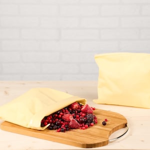 Könnte beinhalten: Zwei gelbe, wiederverwendbare Lebensmittelaufbewahrungsbeutel, einer offen und gefüllt mit gemischten Beeren, darunter Erdbeeren, Himbeeren und Blaubeeren, auf einem Holzschneidebrett. Der andere Beutel ist geschlossen. Eine Glaskaraffe und ein Glas befinden sich im Hintergrund.