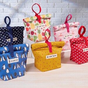 May include: Five reusable lunch bags with different patterns. The bags are made of fabric and have a drawstring closure. The patterns include polka dots, penguins, fruit, flamingos, and hearts. The bags are all labeled with the text "coole Tasche" which is German for "cool bag".