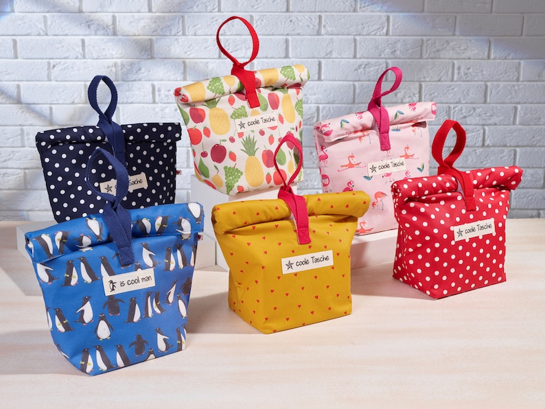 May include: Five reusable lunch bags with different patterns. The bags are made of fabric and have a drawstring closure. The patterns include polka dots, penguins, fruit, flamingos, and hearts. The bags are all labeled with the text "coole Tasche" which is German for "cool bag".