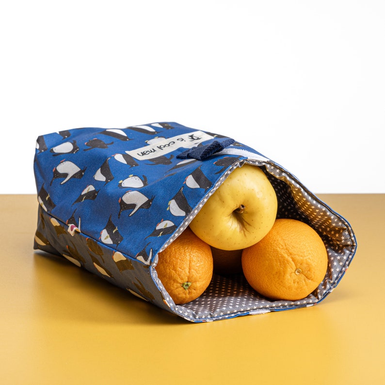 May include: A blue reusable food bag with a penguin pattern and the text "New KooL & Co." printed on the front. The bag is filled with fruit, including apples and oranges.