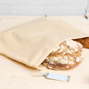Peut inclure: Un sac à pain en coton beige avec une fermeture à cordon, contenant deux pains. Un pain est un pain rond au levain brun foncé, et l'autre est une baguette longue brun clair. Une pomme rouge se trouve sur une surface en bois à côté du sac.
