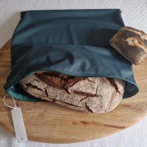 Könnte beinhalten: Ein dunkelgrüner Brotbeutel aus Stoff mit Reißverschluss, der einen großen, krustigen Laib dunkles Brot enthält. Ein kleineres, mit Mohn bestreutes Brötchen liegt außerhalb des Beutels. Ein weißes Kunststoffetikett ist am Reißverschluss befestigt.