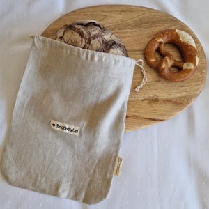Könnte beinhalten: Ein beiger Brotbeutel aus Leinen mit dem Text "Brotbeutel" auf einem kleinen Etikett. Ein Brotlaib ist teilweise in der Tasche zu sehen. Ein Holzschneidebrett hält eine Brezel.