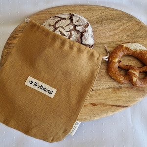 Könnte beinhalten: Ein beiger Brotbeutel mit dem Text "Brotbeutel" auf einem weißen Etikett, der ein Brot enthält. Ein Brezel und ein rundes Holzschneidebrett sind ebenfalls zu sehen.