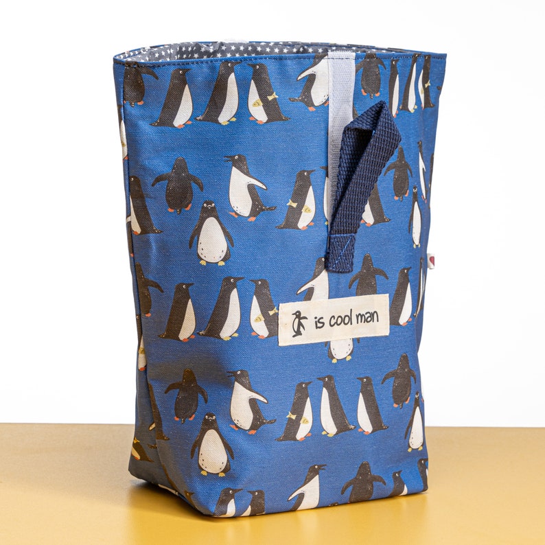 May include: A blue reusable shopping bag with a penguin pattern. The bag has a black handle and a white tag that says "is cool man".