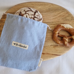 Könnte beinhalten: Ein hellblaues Brotbeutel mit dem Text "Brotbeutel" auf einem weißen Etikett, das ein Brot enthält. Ein Brezel und ein rundes Holzschneidebrett sind ebenfalls zu sehen.