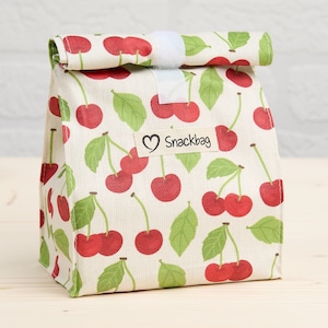 Könnte beinhalten: Eine wiederverwendbare Snacktasche mit einem Muster aus roten Kirschen und grünen Blättern auf cremefarbenem Hintergrund. Die Tasche hat eine gerollte Oberseite, die mit einem weißen Klettverschluss befestigt ist. Ein Etikett mit einem Herz und dem Wort "Snackbag" ist angebracht.